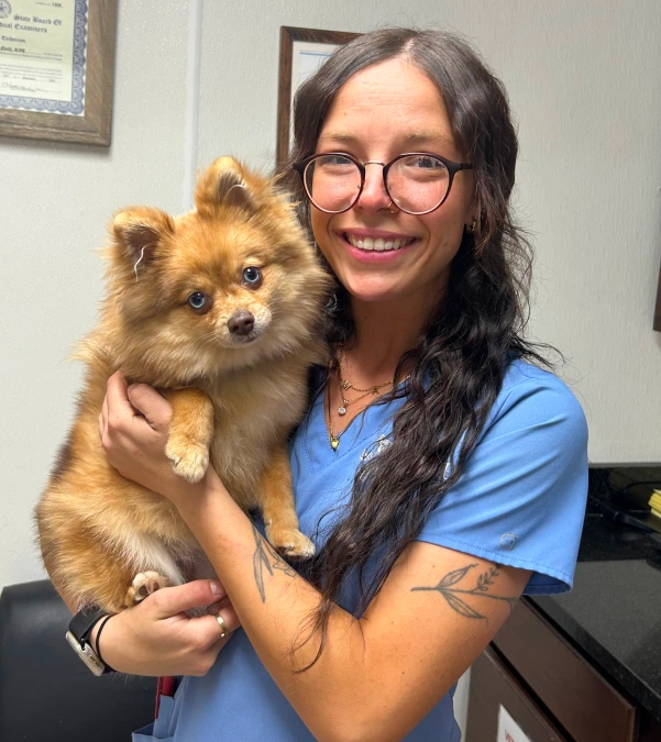 A smiling woman with long dark hair, glasses, and a leaf tattoo holds a fluffy orange Pomeranian dog, wearing blue scrubs in an office with a framed certificate on the wall.