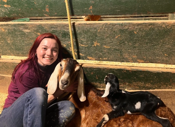 A smiling woman with red hair crouches beside a brown goat with long floppy ears while a black and white baby goat lies nearby, inside a wooden stall with straw bedding.